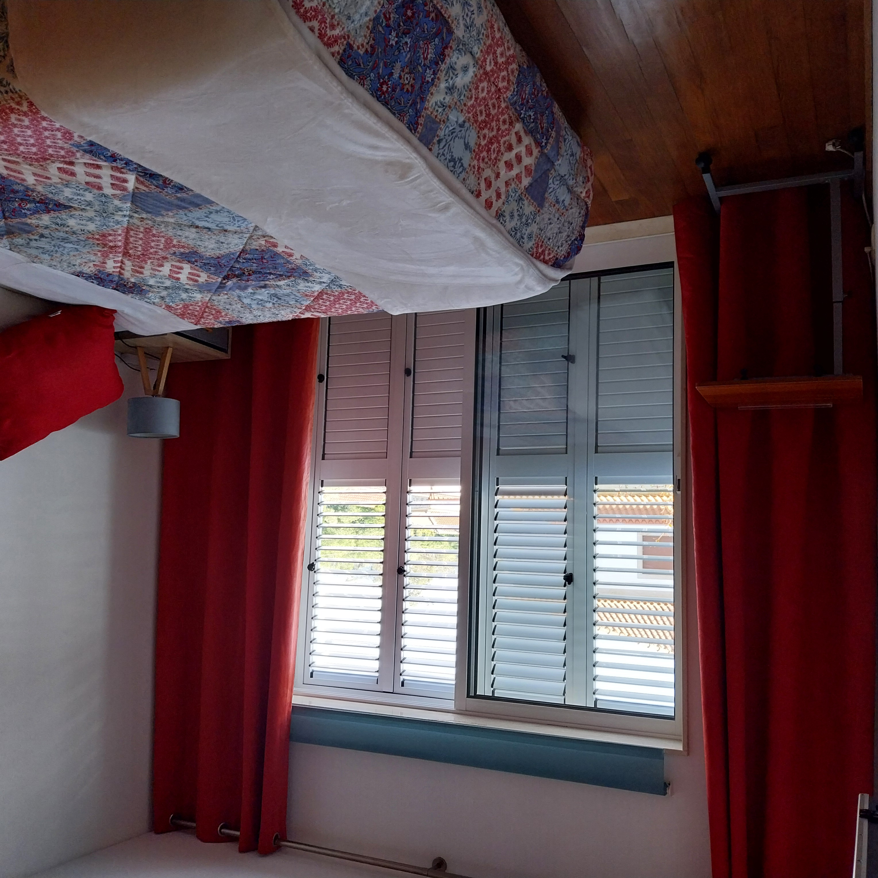 Quarto Duplo com Varanda com portadas brancas, cortinado vermelho e cama de casal com colcha em padrão